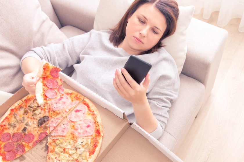 Junge Frau liegt gestresst mit Pizza auf dem Sofa.